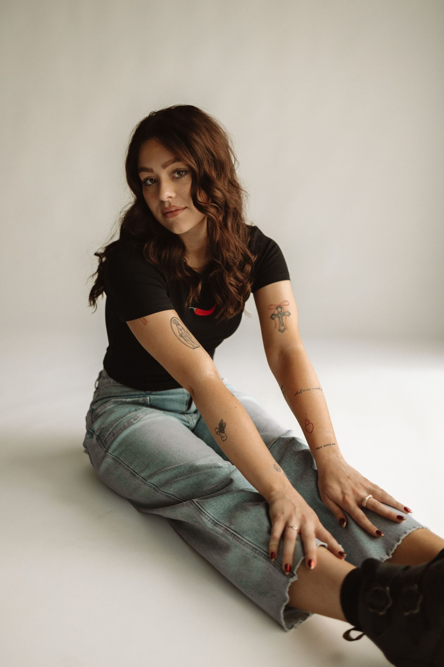 Person in black t-shirt and distressed jeans sitting on white floor with visible arm tattoos.