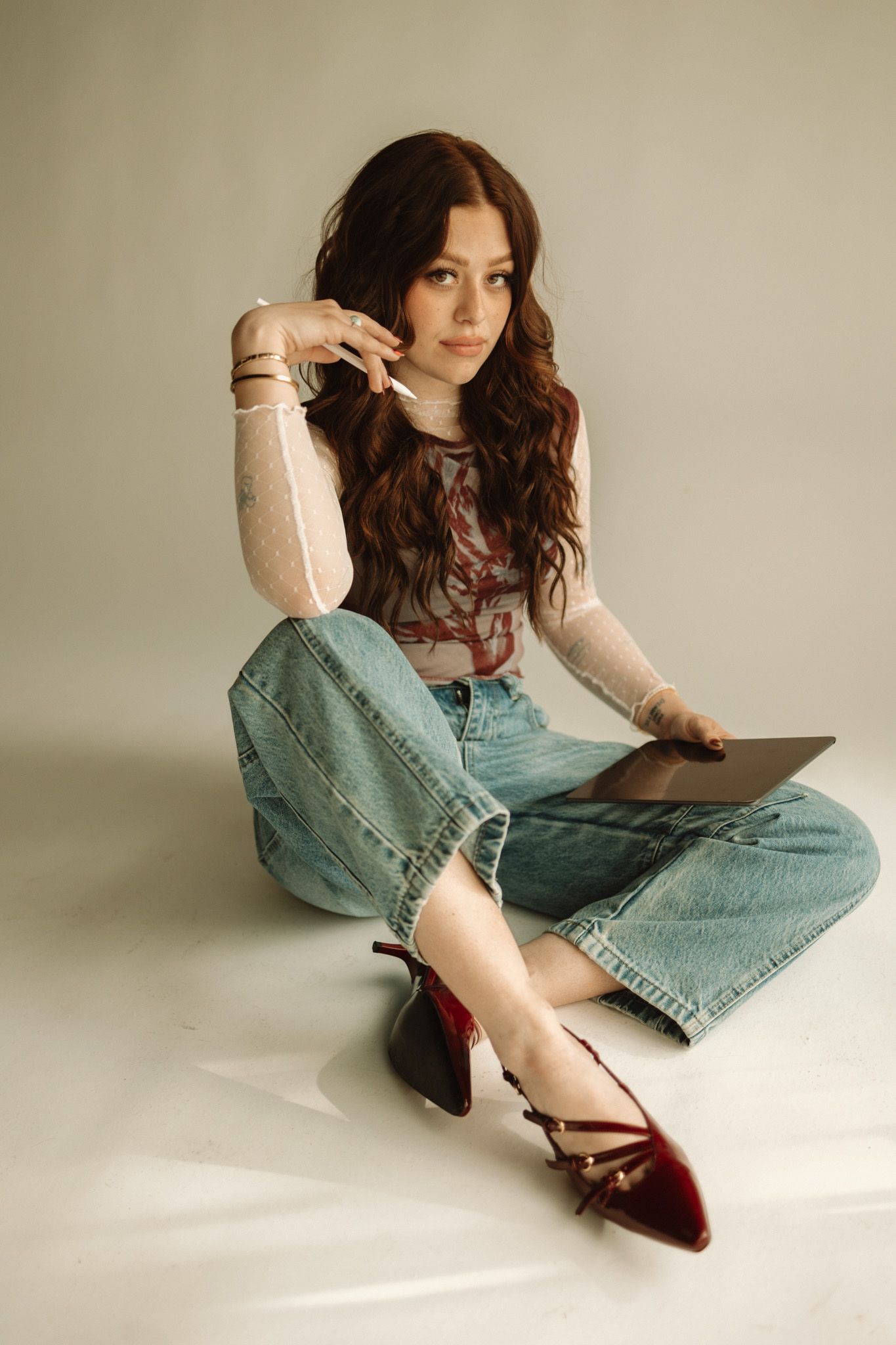 Young woman in polka dot top and baggy jeans sits casually on floor wearing burgundy strappy flats against beige backdrop.