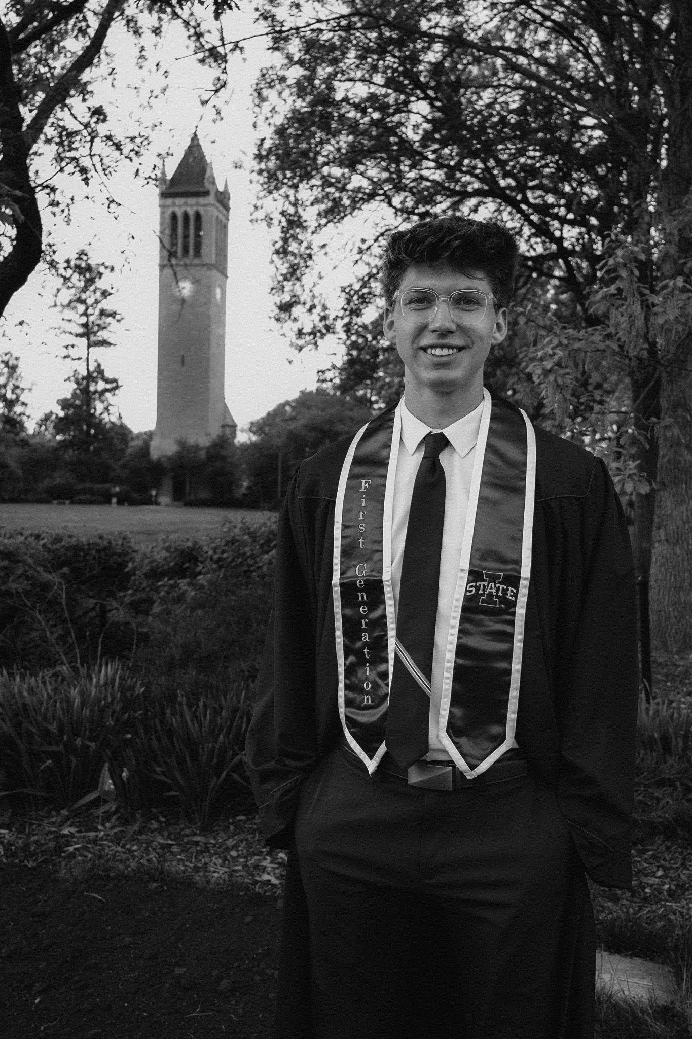 A graduate in cap and gown poses in front of a tall bell tower on a university campus.