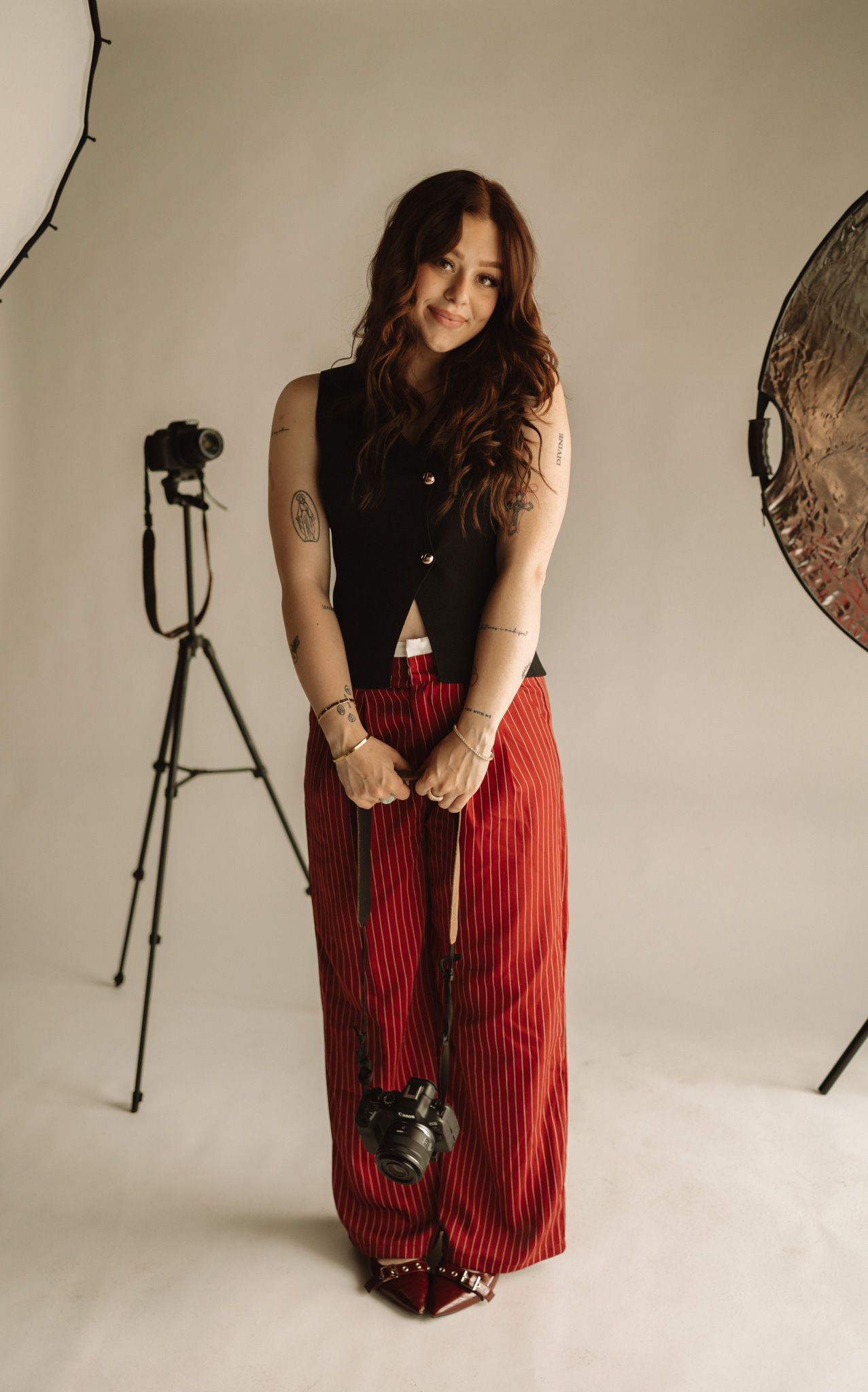 Studio photography setup with model wearing black top and red striped pants posing between lighting equipment.