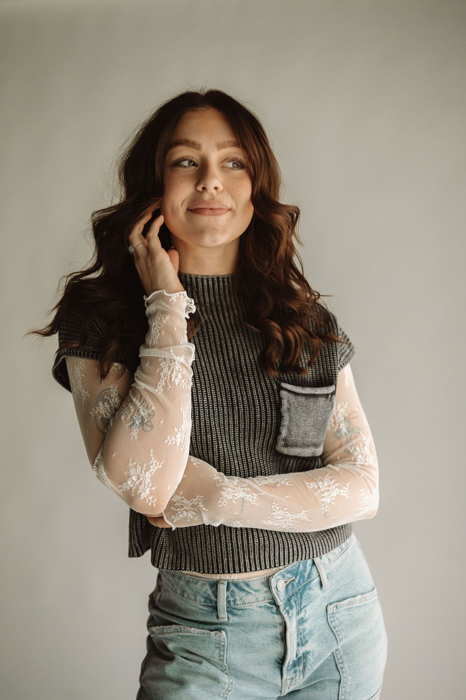 Woman wearing a sheer long sleeve top with dark vest and light denim jeans poses against neutral wall.