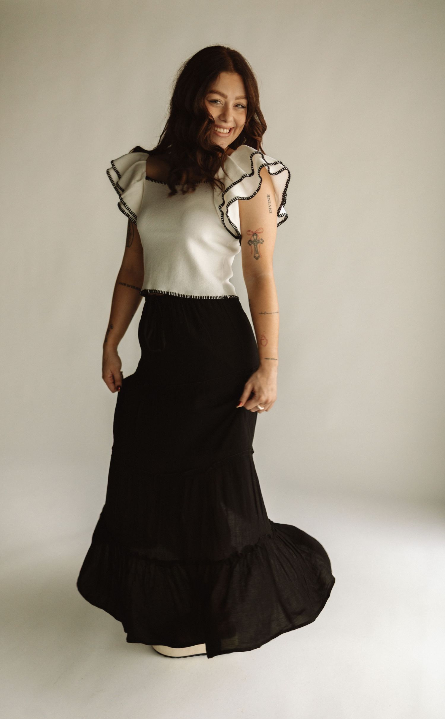 A woman in a white ruffled top and flowing black maxi skirt poses against a neutral backdrop in a studio setting.