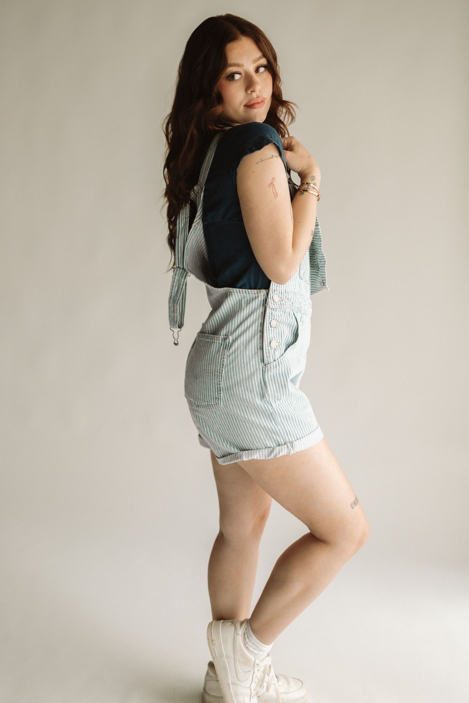 A person in a black tank top and light blue striped overalls poses against a neutral background wearing white sneakers.