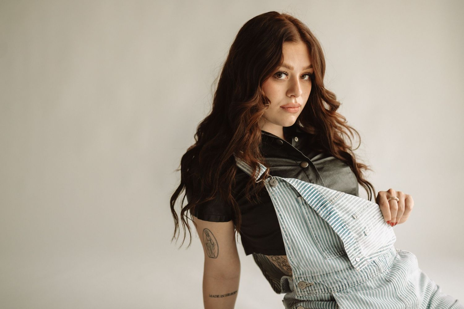 A young woman in blue denim overalls and black shirt poses against a neutral background with flowing dark hair.