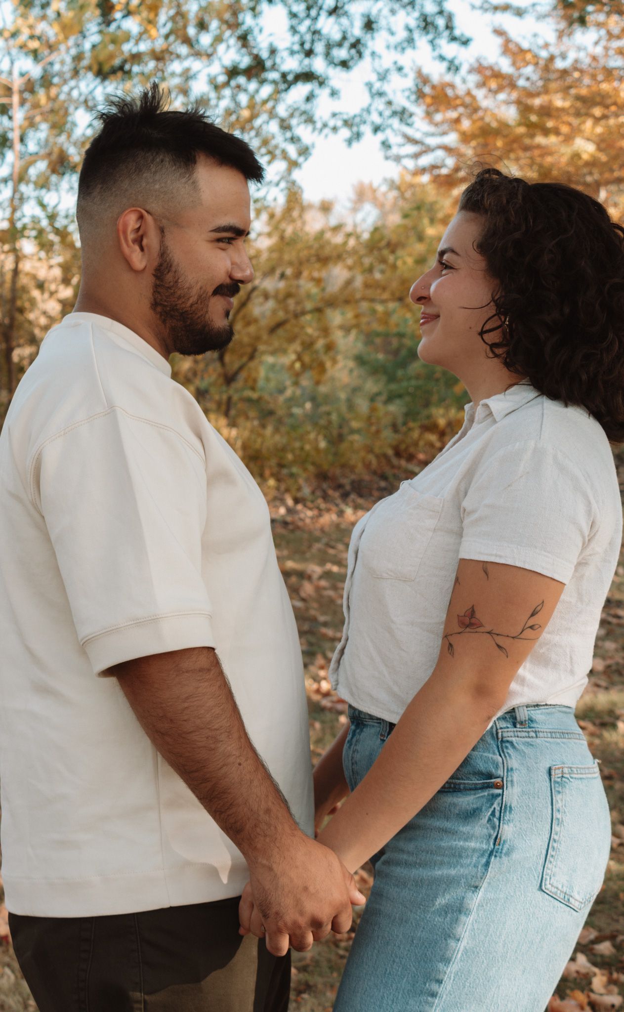 A couple holds hands while gazing at each other lovingly against an autumn backdrop with golden foliage.