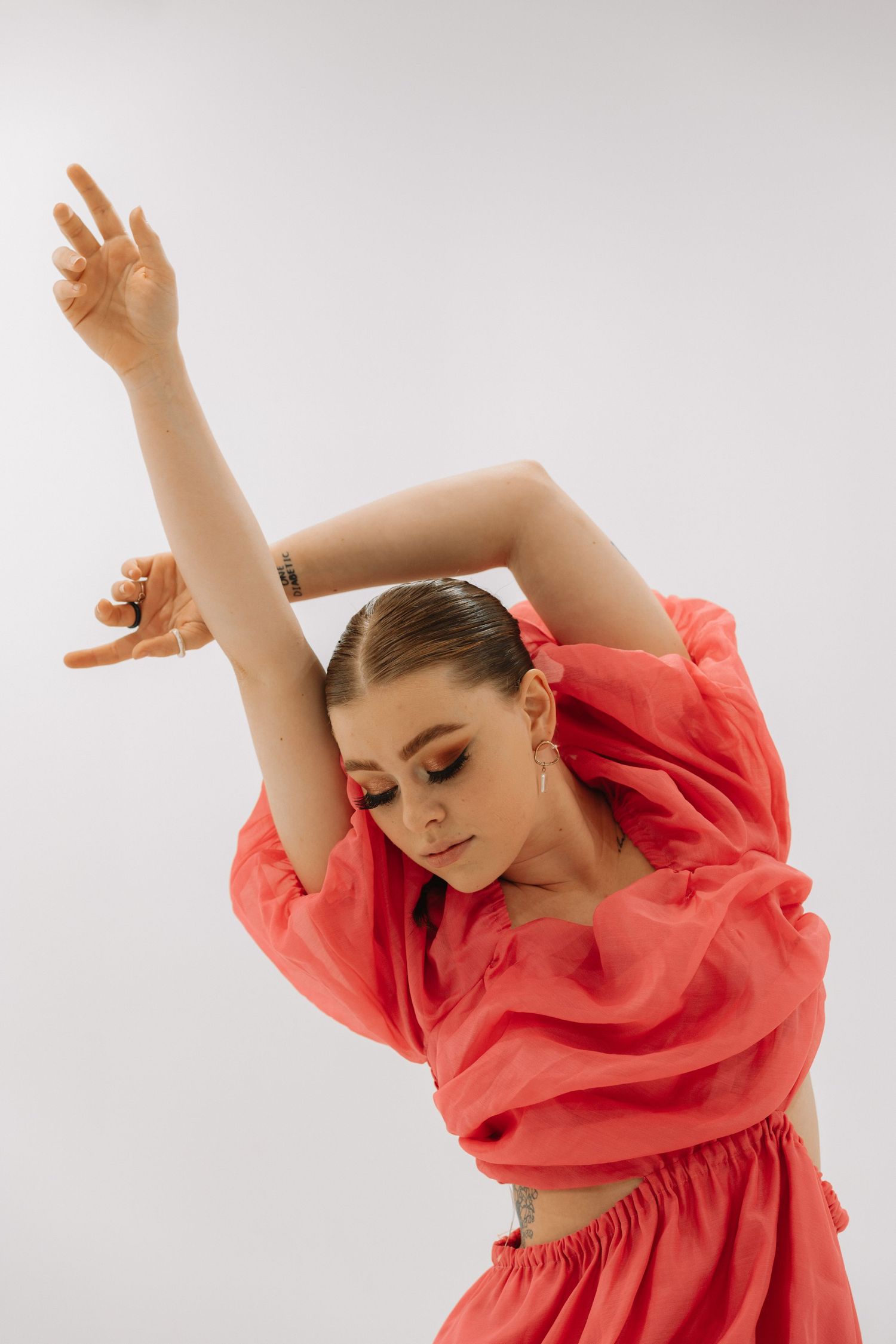Dancer in flowing coral dress performs graceful movement with raised arms against white background.
