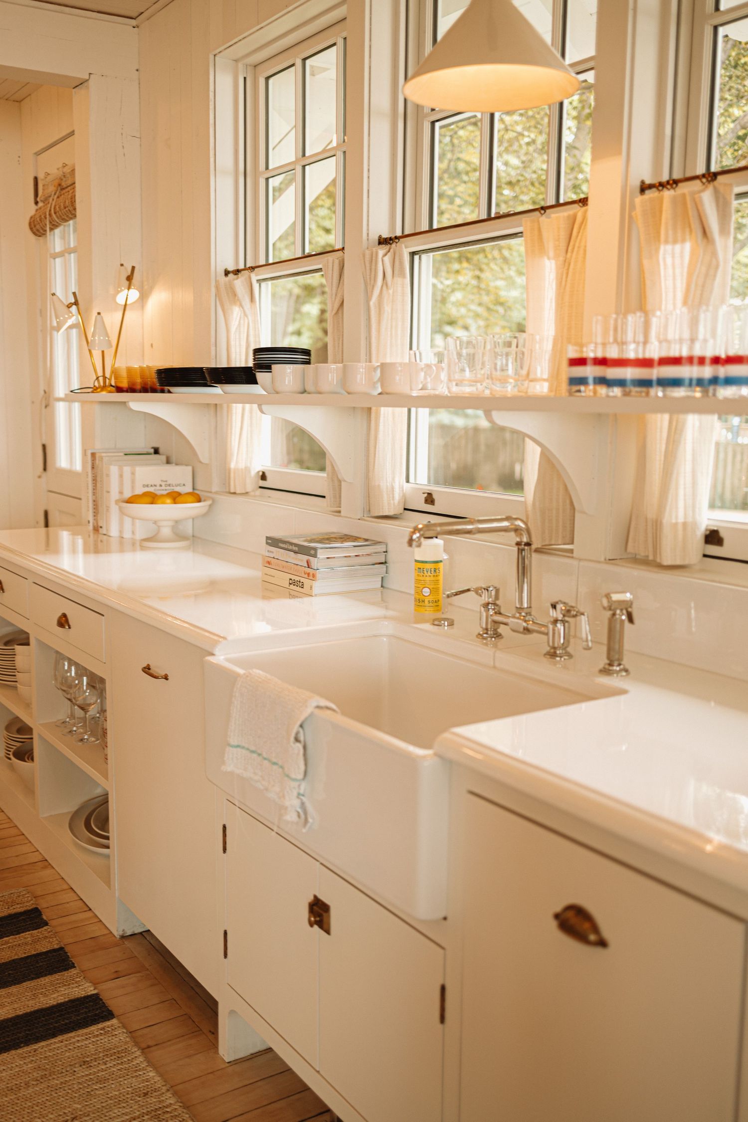 Modern white kitchen with farmhouse sinks, white cabinets, and pendant lighting in bright natural light from windows.