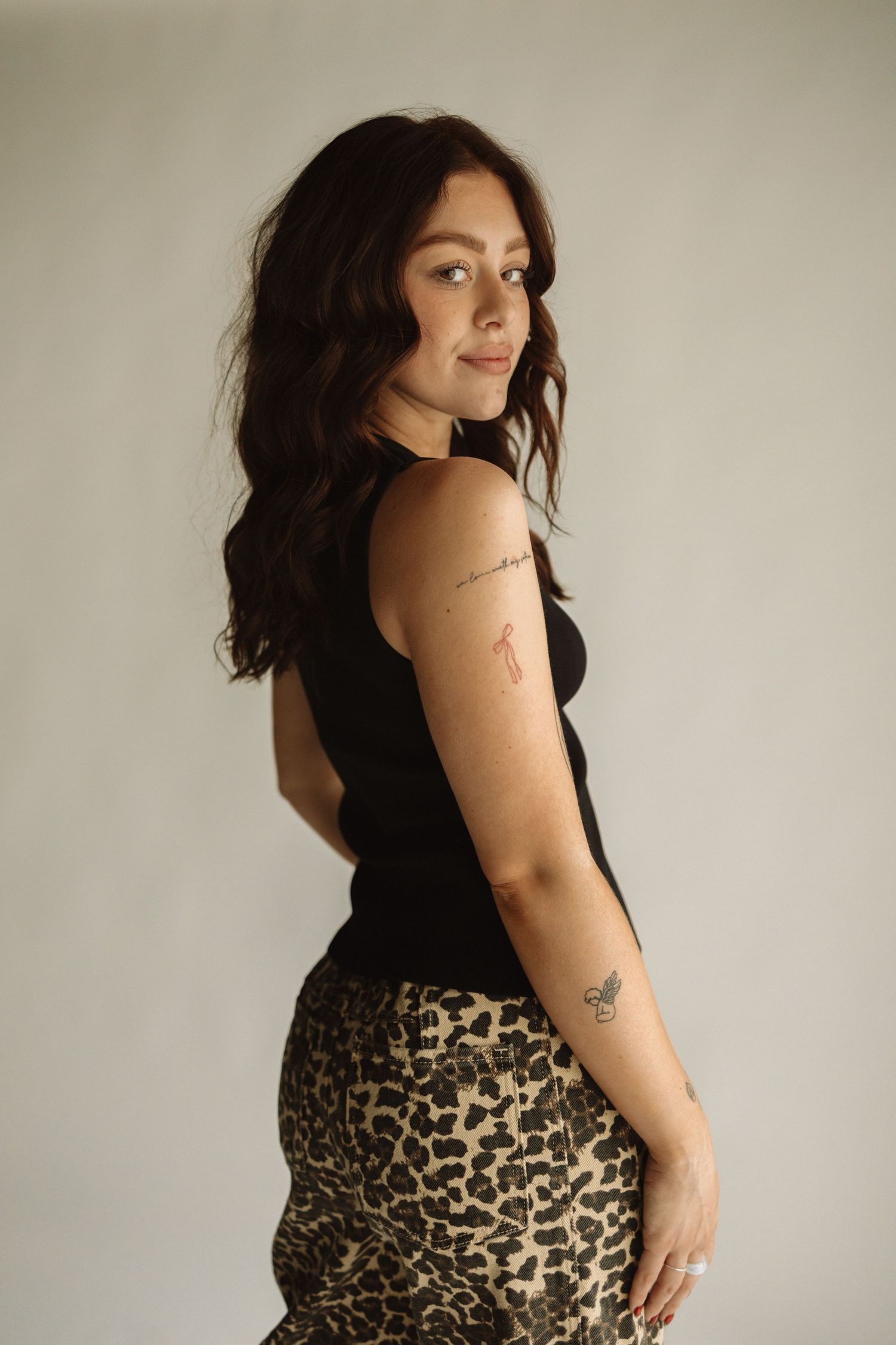 A woman in a black tank top and leopard print skirt poses against a neutral wall, showing minimalist tattoos on her arm.