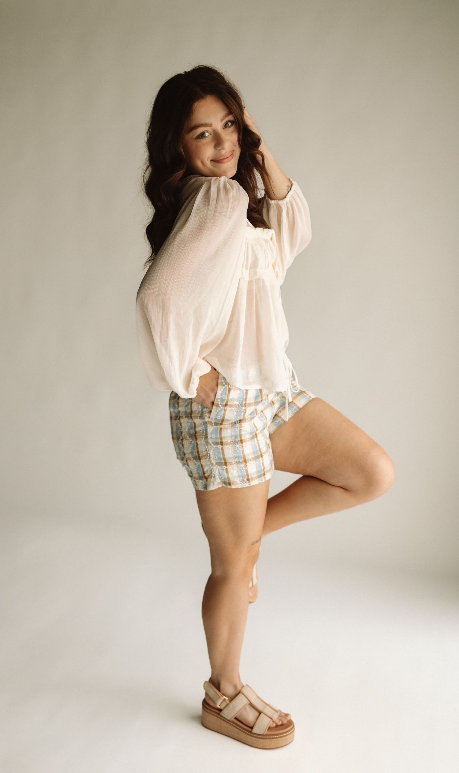 Fashion model wearing cream colored top and checkered shorts poses elegantly in studio setting with white background.