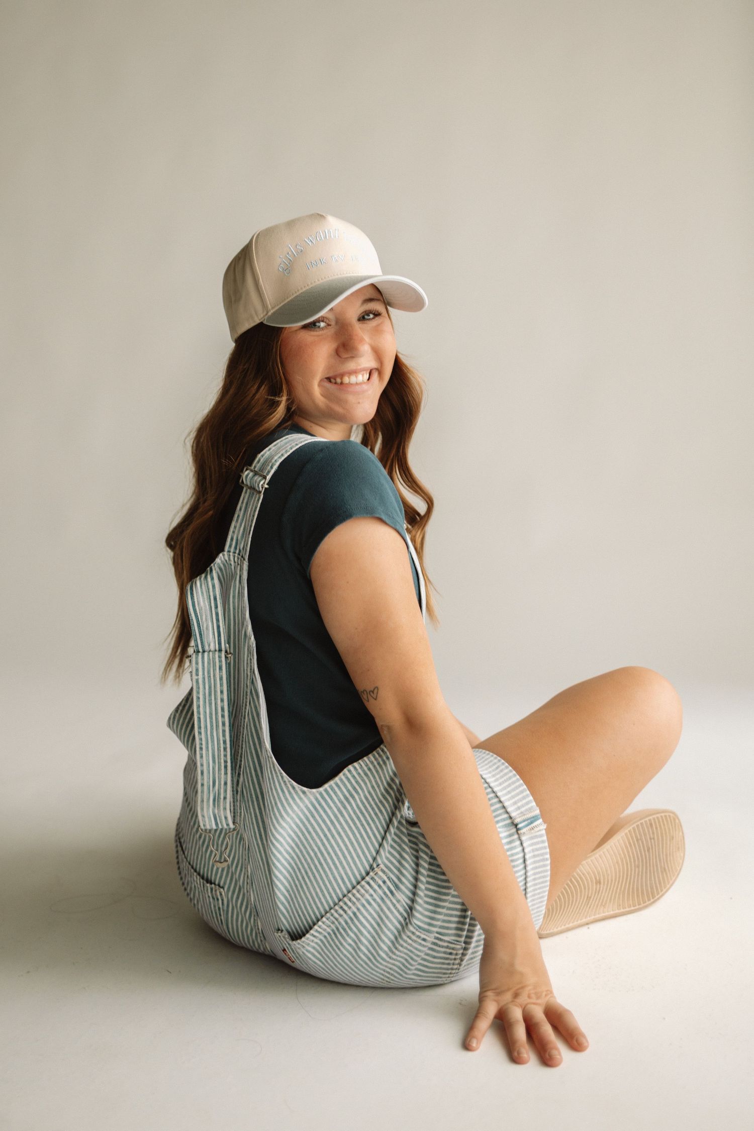 A figure wearing a striped romper and baseball cap reclines with a playful smile against a neutral studio backdrop.