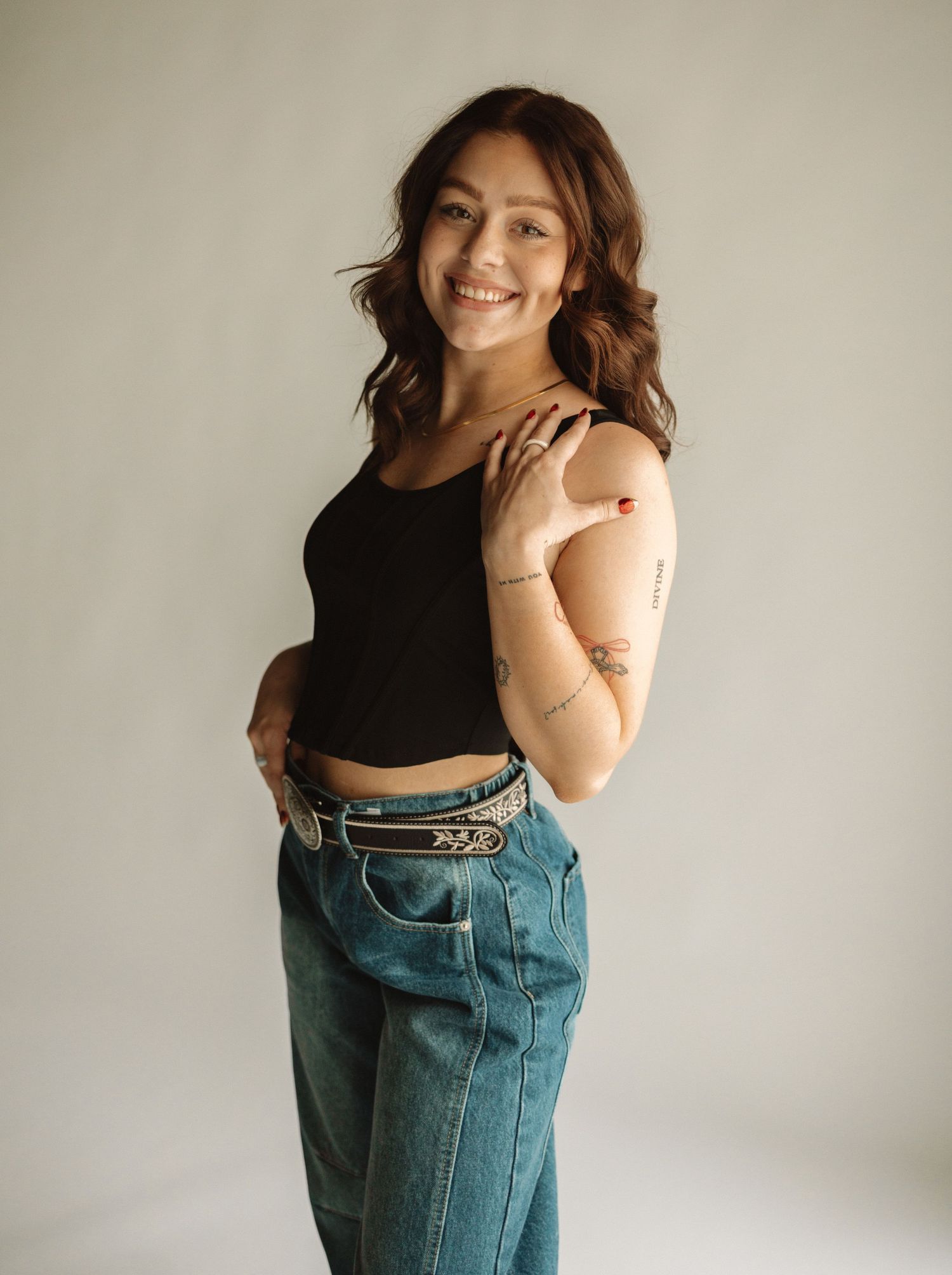 A person in a black crop top and high-waisted jeans poses against a neutral background with natural hair and smile.