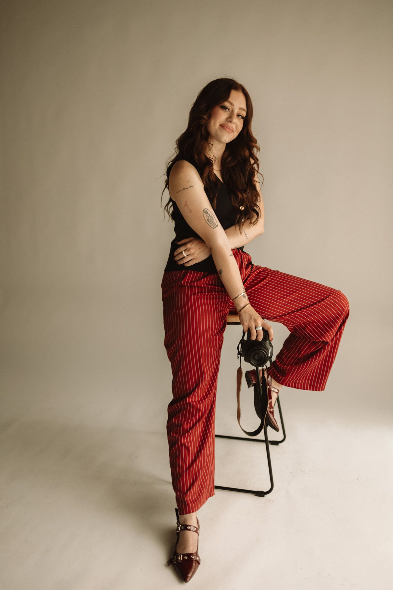 A person in black top and red striped wide leg pants poses on stool against neutral backdrop for fashion shot.