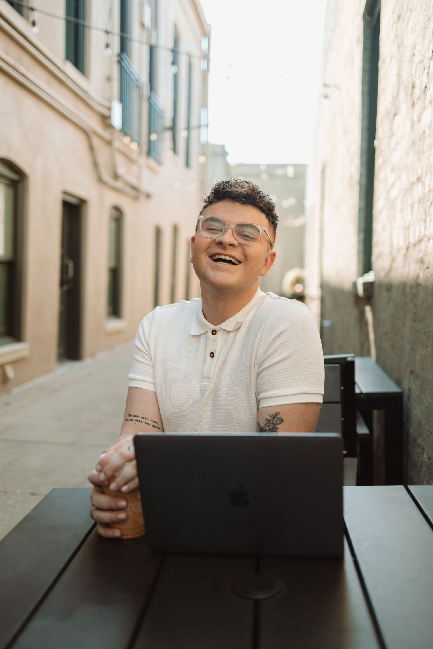 Person sitting outdoors at cafe table with laptop and beverage enjoying sunny day with joyful expression.