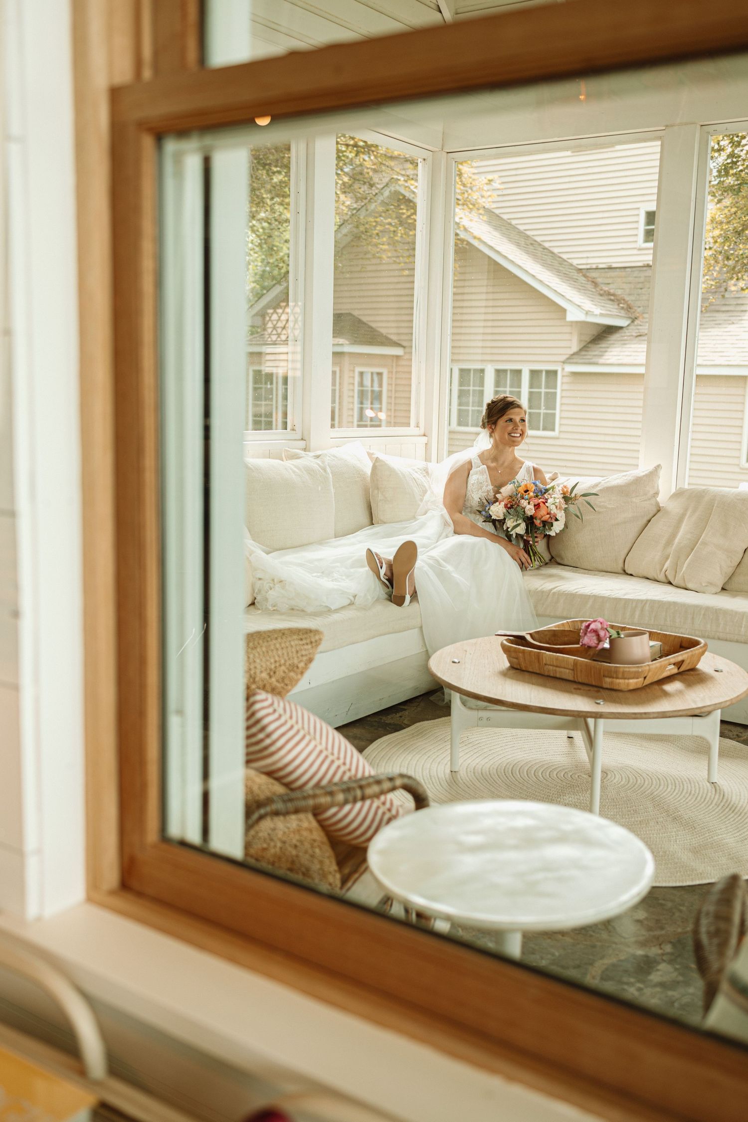 View through window of bright sunlit porch with white sectional sofa, wooden coffee table, and pink floral bouquet.