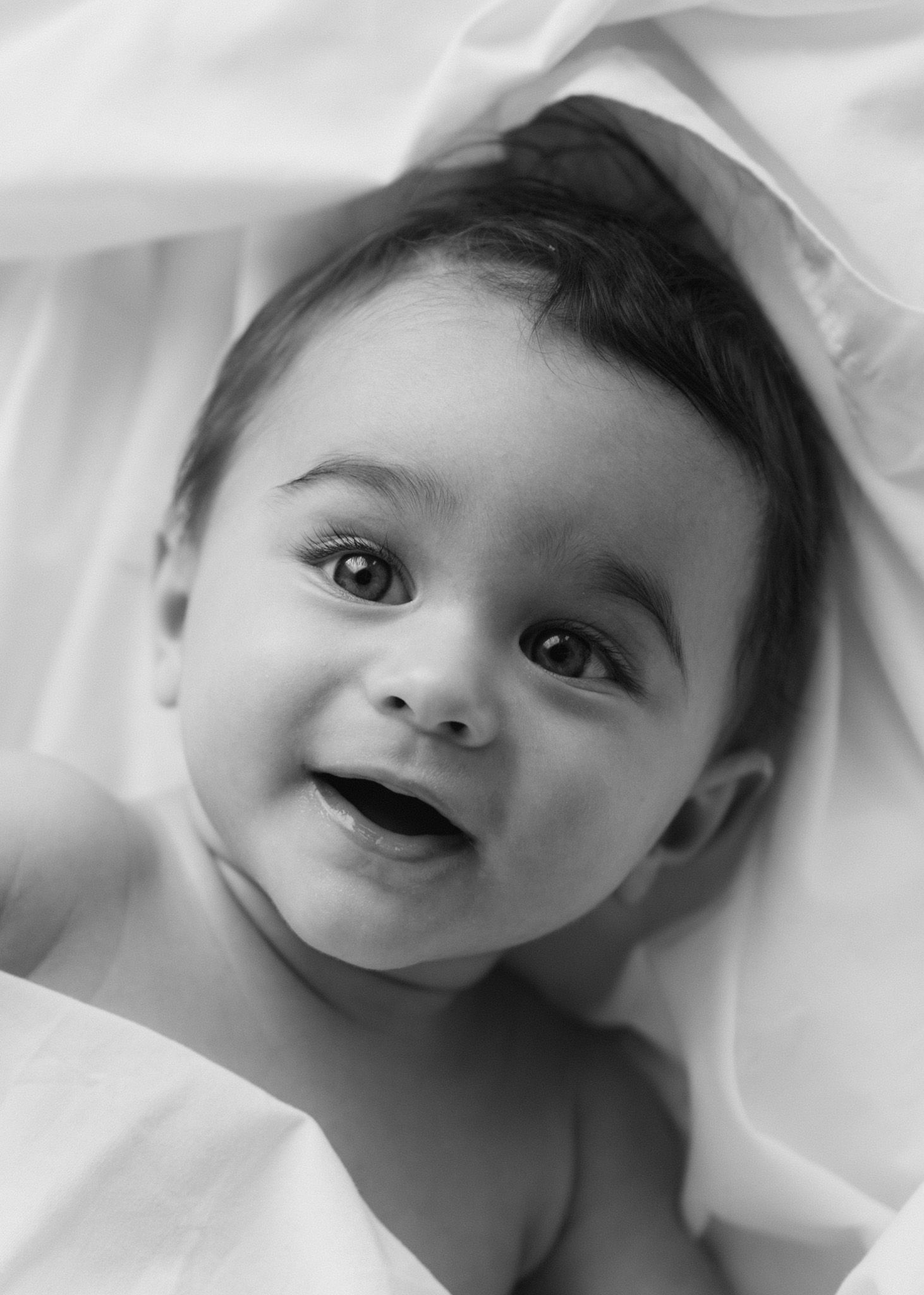 Black and white close-up of a baby smiling while lying on white bedding.
