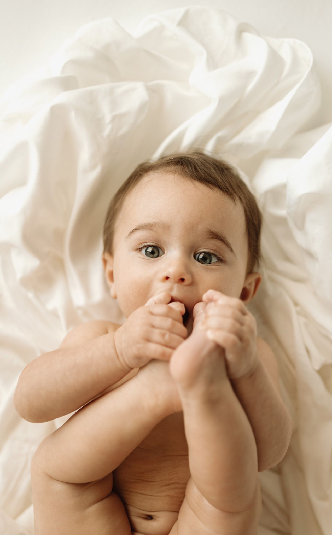 A baby lies on white bedding while playfully grabbing their own feet in a series of adorable poses.