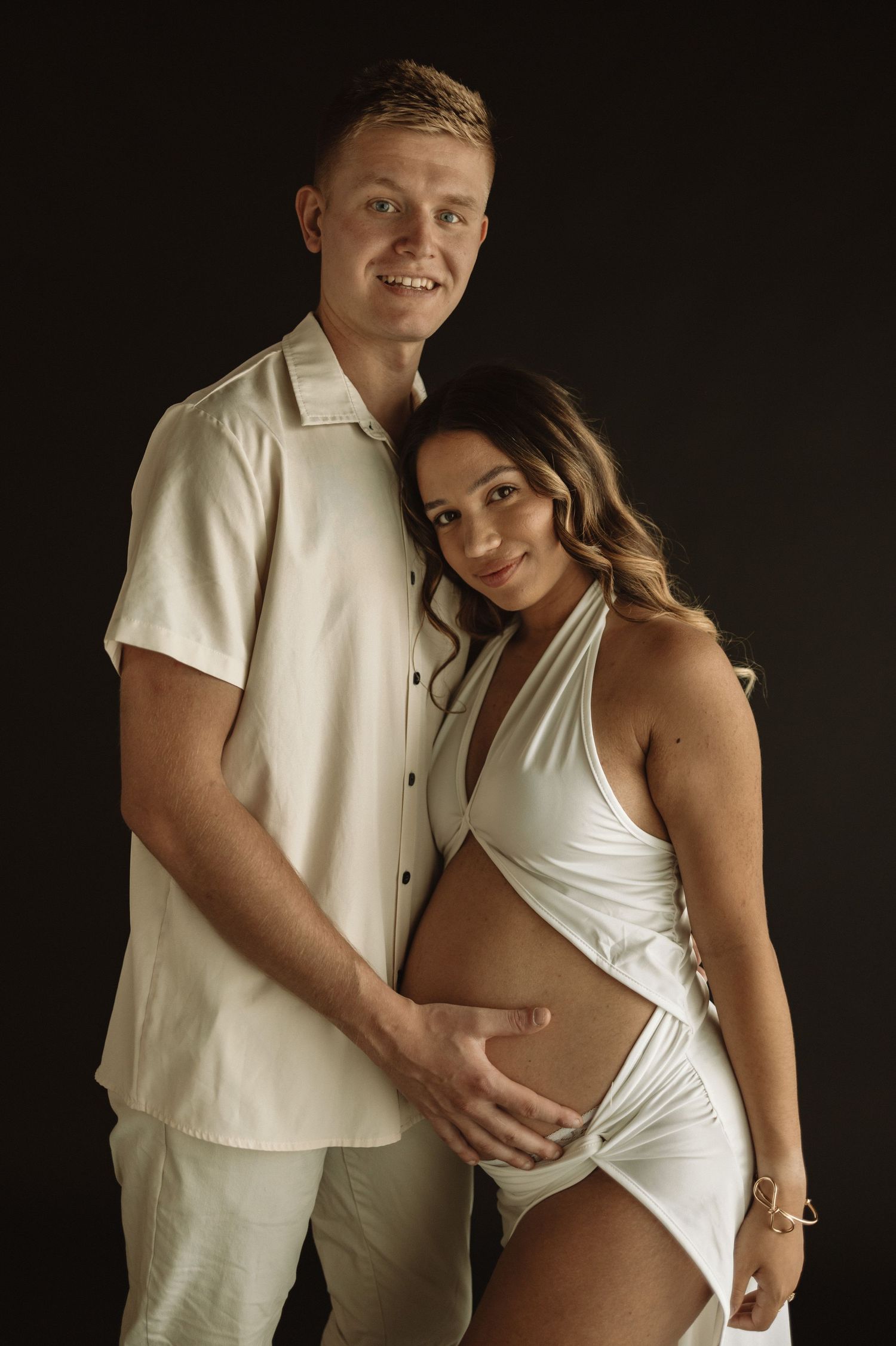 Four intimate maternity photos showing a couple in white attire posing together against a dark background.