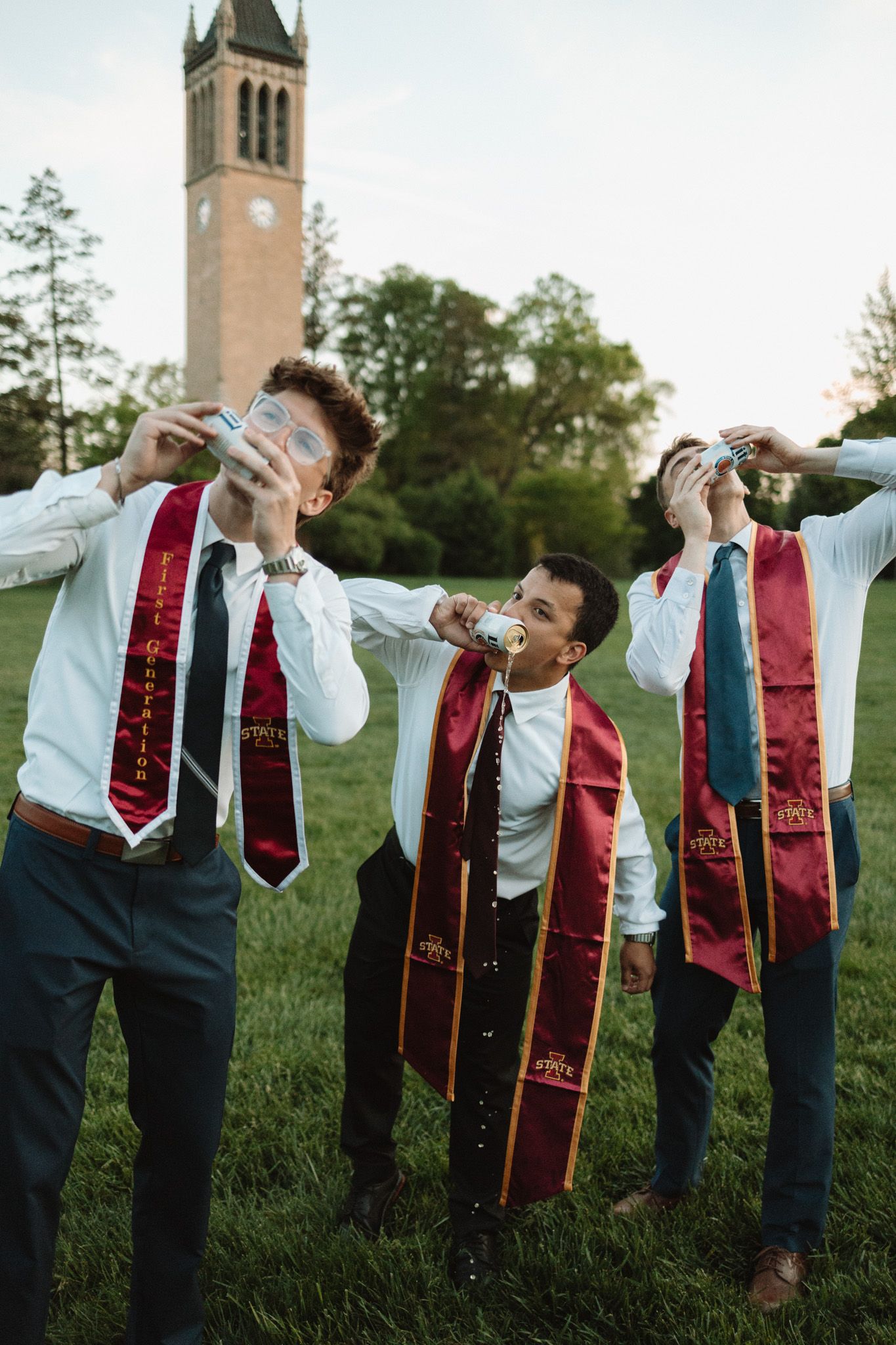 Graduates in dress shirts and red stoles line up to form a playful human domino effect on a grassy field.