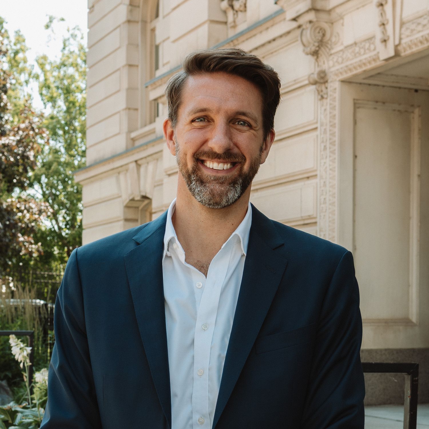 Professional headshot of someone in a navy blazer and white shirt smiling outdoors near a building with ornate architecture.