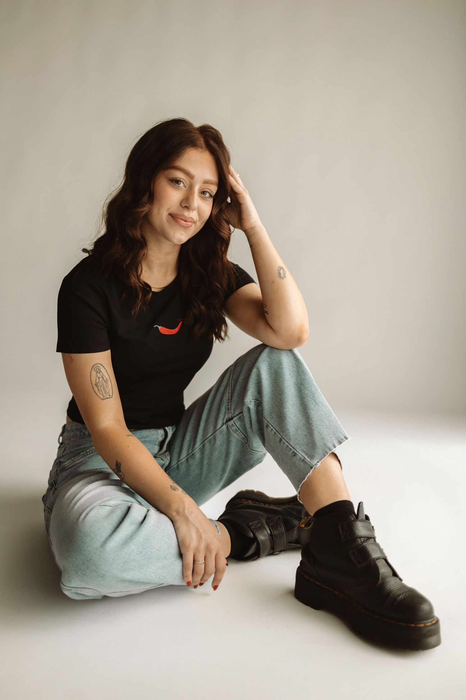 Portrait of someone in a casual black t-shirt and light wash jeans sitting against a white backdrop wearing chunky boots.