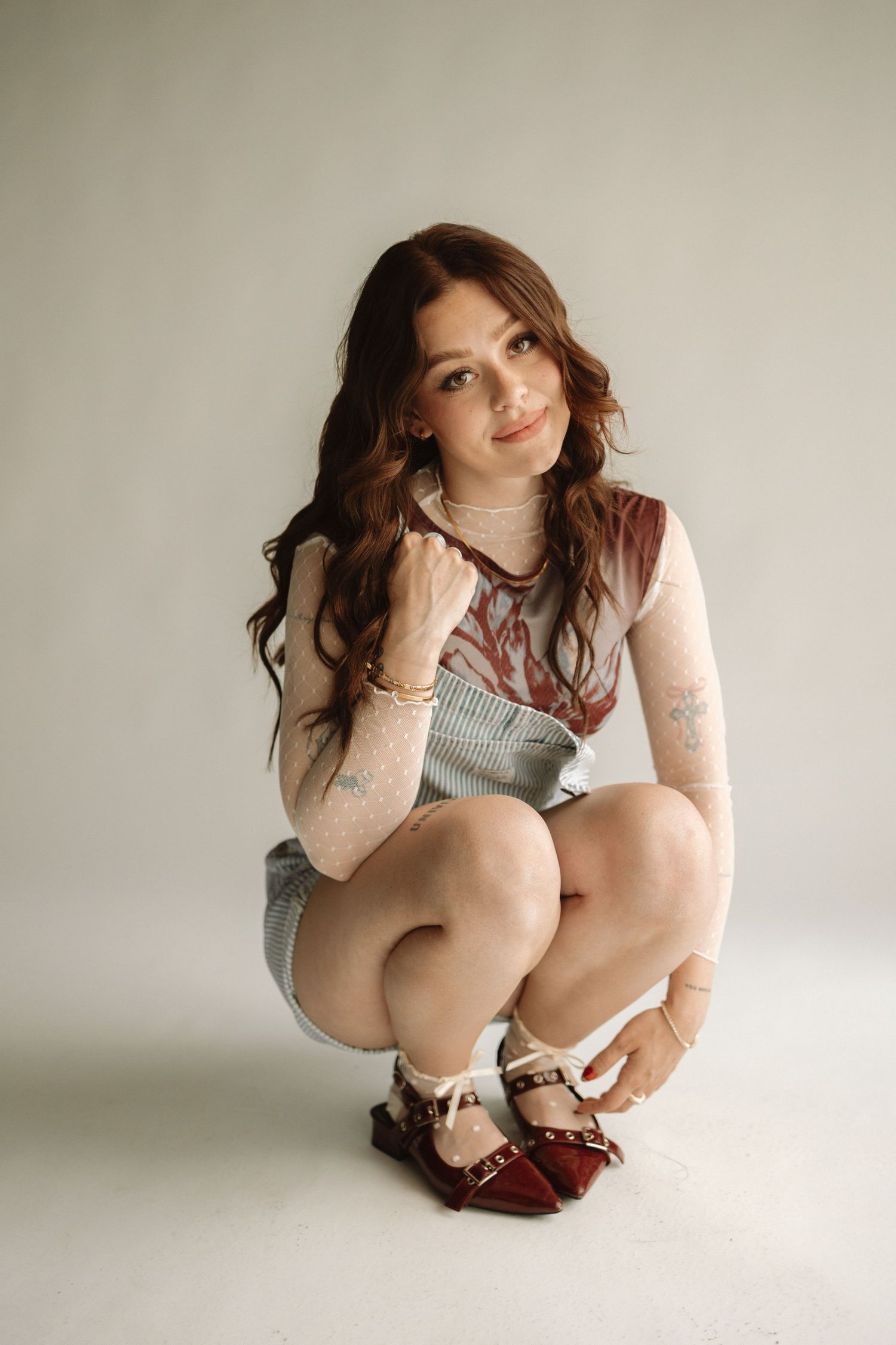 A person in a light mesh top and denim shorts crouches while wearing burgundy oxford shoes against a neutral backdrop.