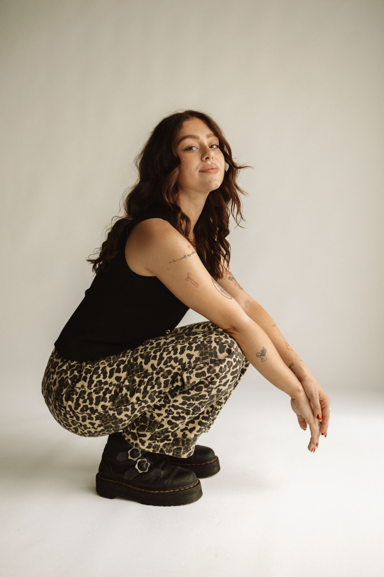 Person wearing black tank top and leopard print pants crouching in studio setting against white background with combat boots.