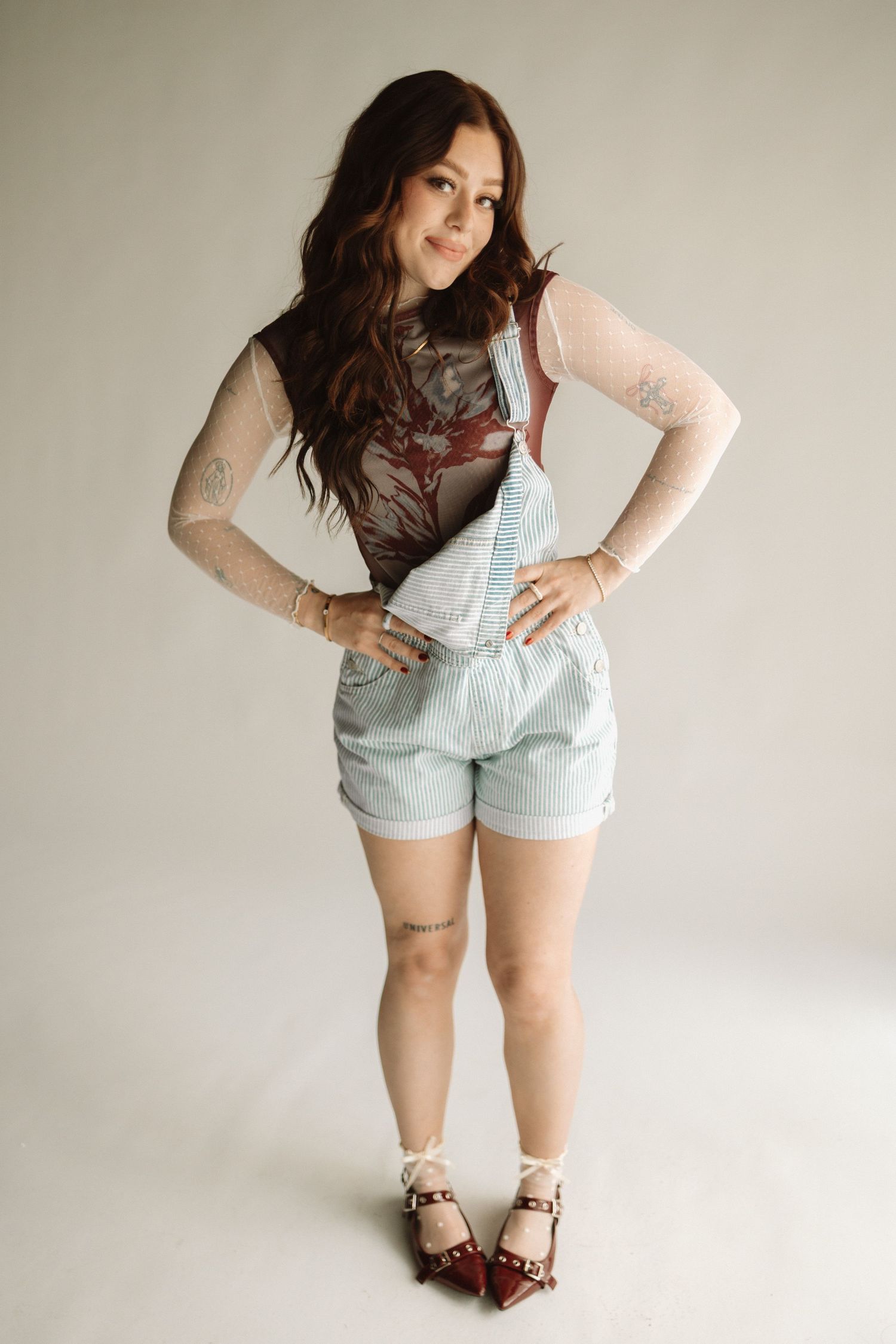 Model wearing light denim overalls with sheer sleeves and burgundy shoes poses against neutral background.