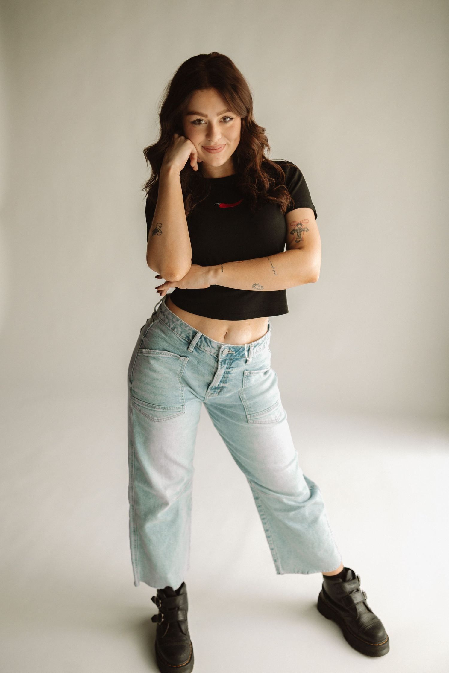 Studio fashion shot featuring model in casual black top, faded jeans and dark boots in thoughtful posed position.