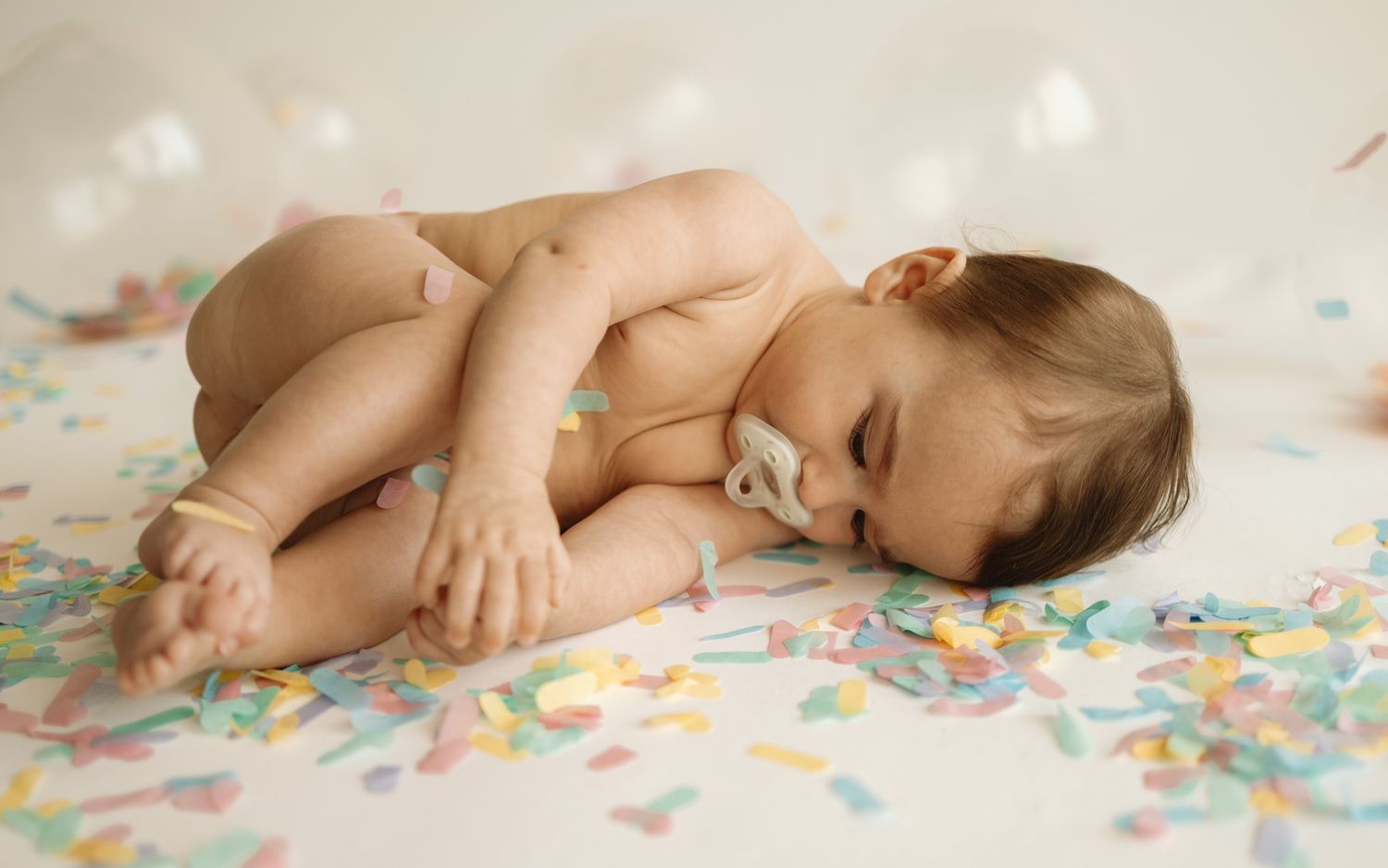 A sleeping baby curled up on colorful confetti-covered bedding.