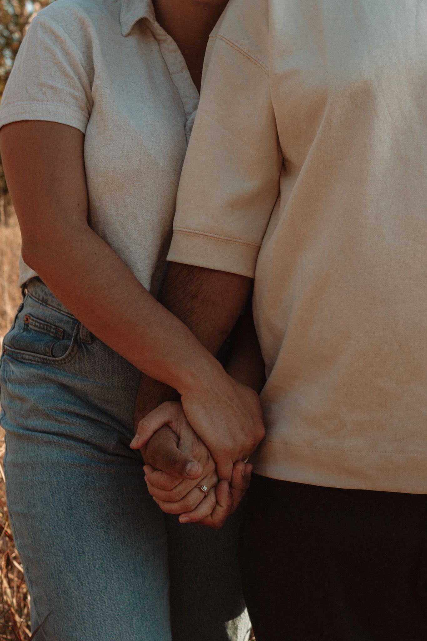 A couple holding hands while wearing blue jeans and casual attire, photographed from the waist down in warm sunlight.