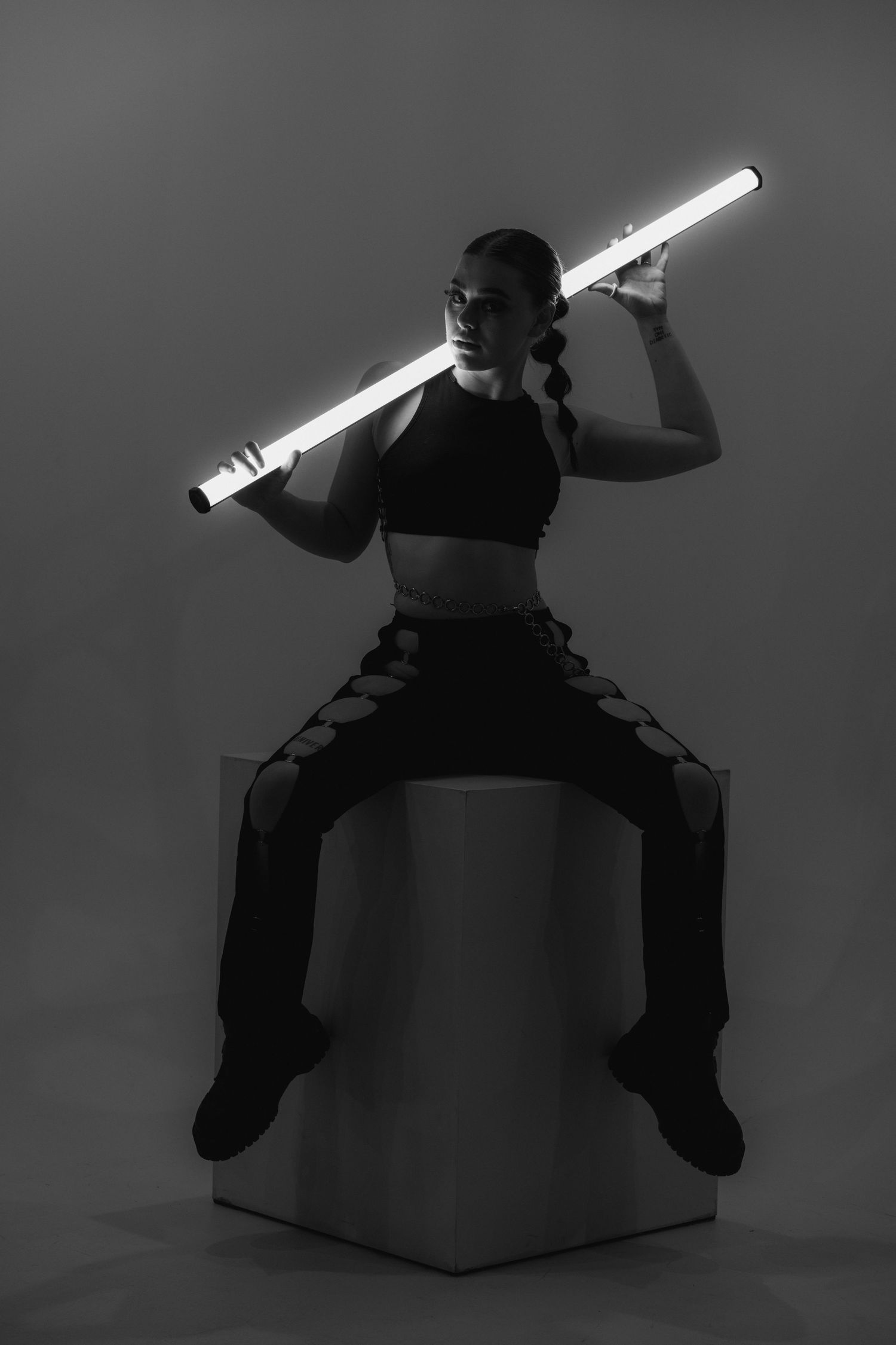 Dramatic black and white photo of a figure in black clothing holding a glowing light saber while posing on a pedestal.