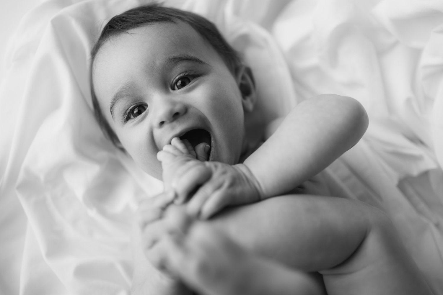 Black and white photo of a laughing baby lying on white bedding.