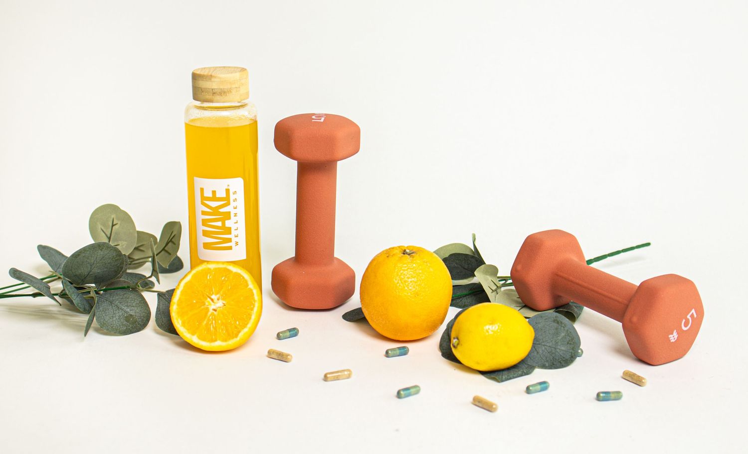 Orange juice bottle and dumbbells arranged with fresh citrus fruits and eucalyptus leaves on white background.