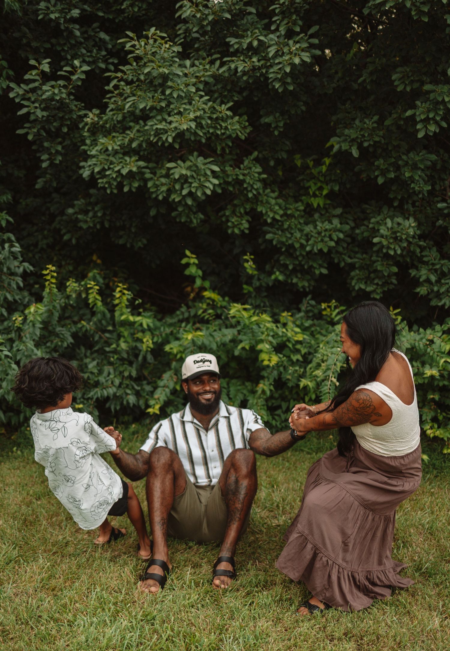 Three people sit casually on grass while relaxing outdoors among green foliage on a sunny day.