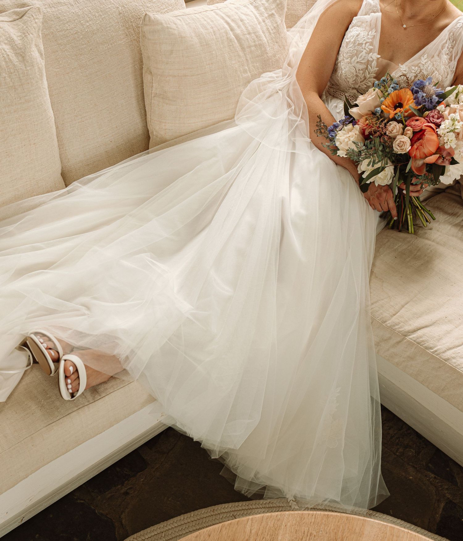 Close-up details of an elegant white wedding dress with tulle fabric and floral bouquet resting on a cream sofa.