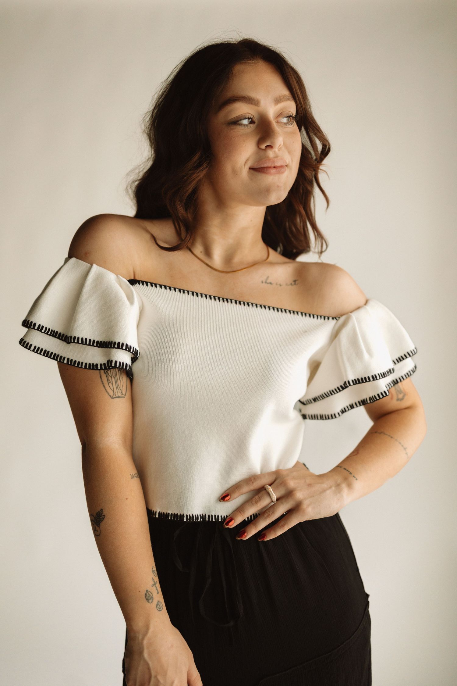 An elegant off-shoulder white top with black trim detail paired with a dark skirt in a minimalist studio setting.