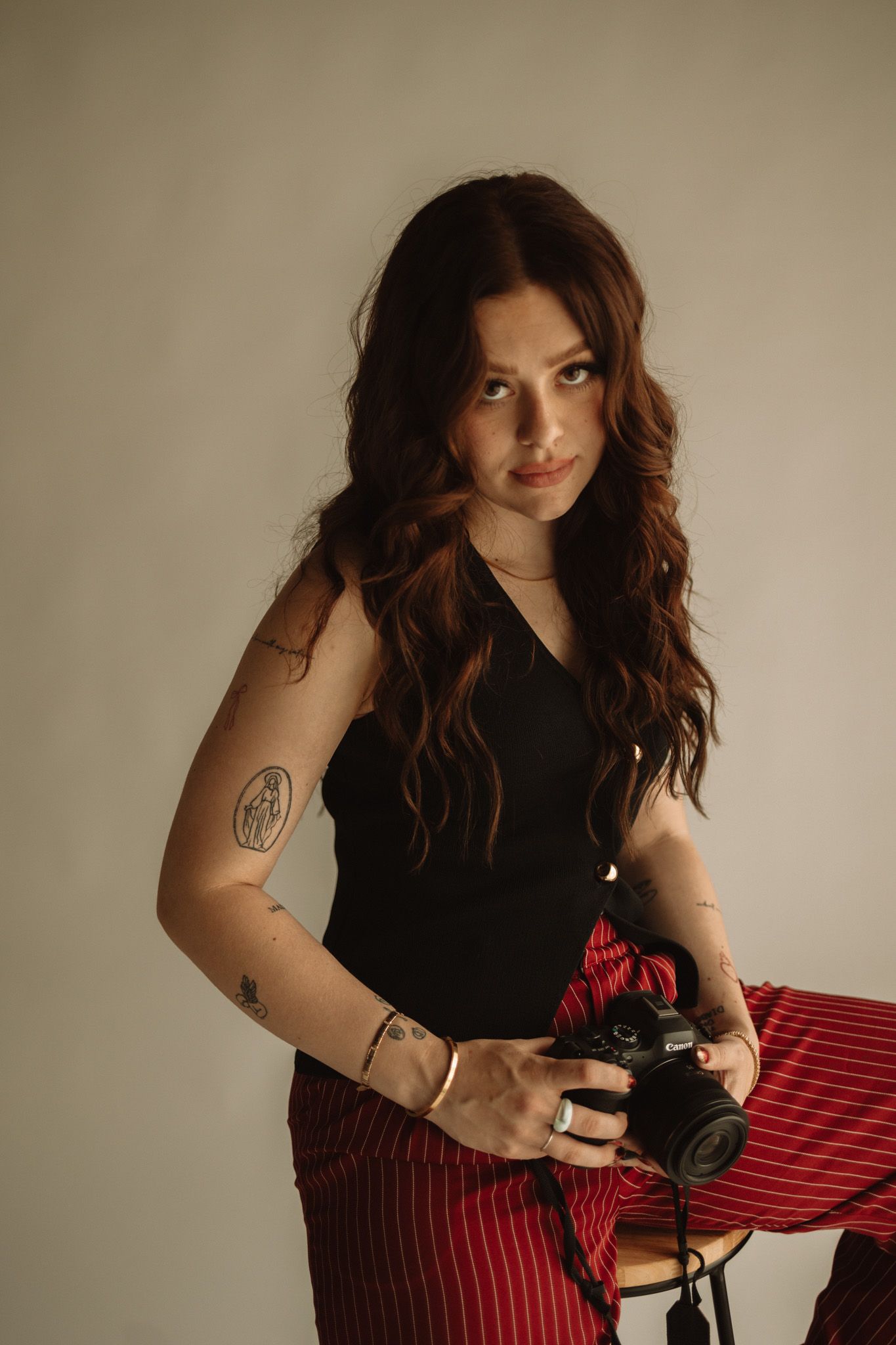 A person in a black top and red striped pants holds a vintage camera against a neutral background.