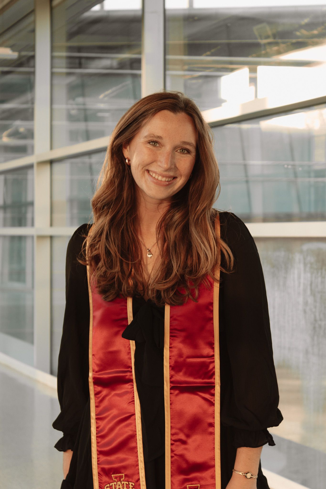 Graduate wearing black graduation gown with red and gold stole stands in bright modern hallway for graduation photo.