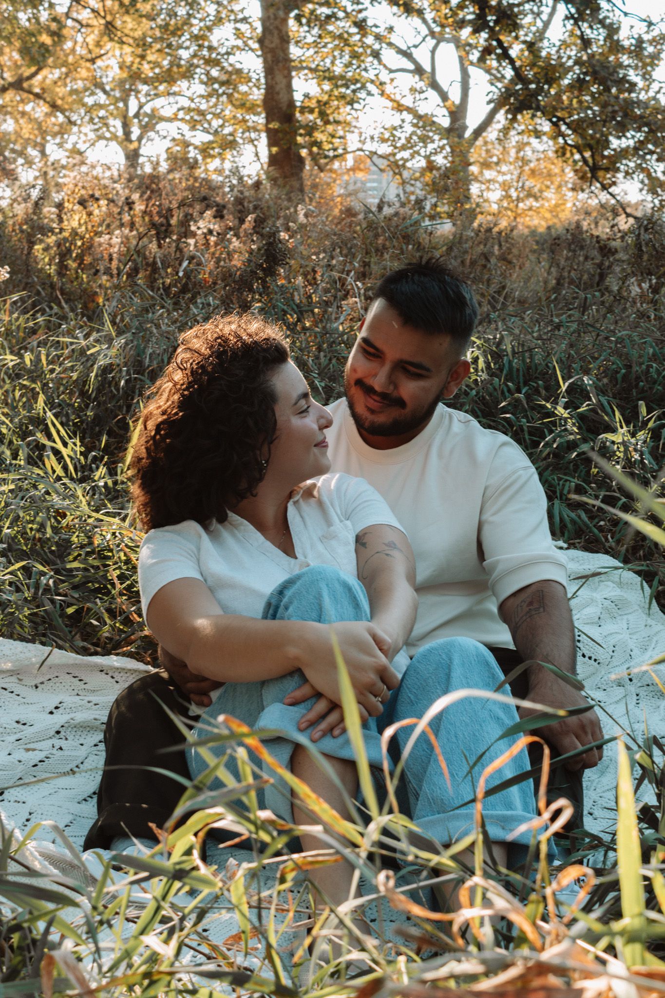 A couple relaxes together on a white blanket among grass and foliage during a romantic outdoor photoshoot.