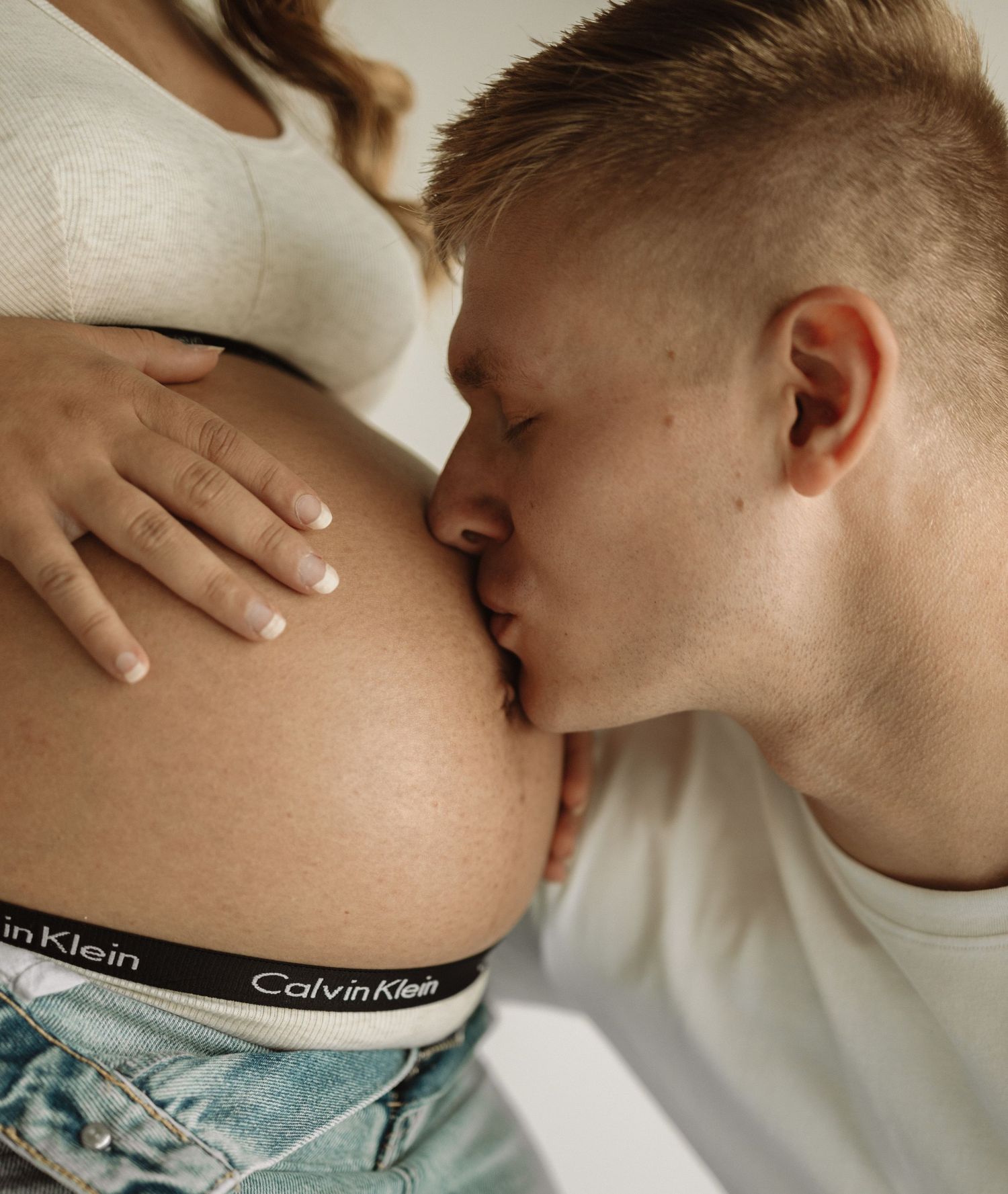Intimate close-up photo of someone kissing a pregnant belly exposed above Calvin Klein underwear and denim.