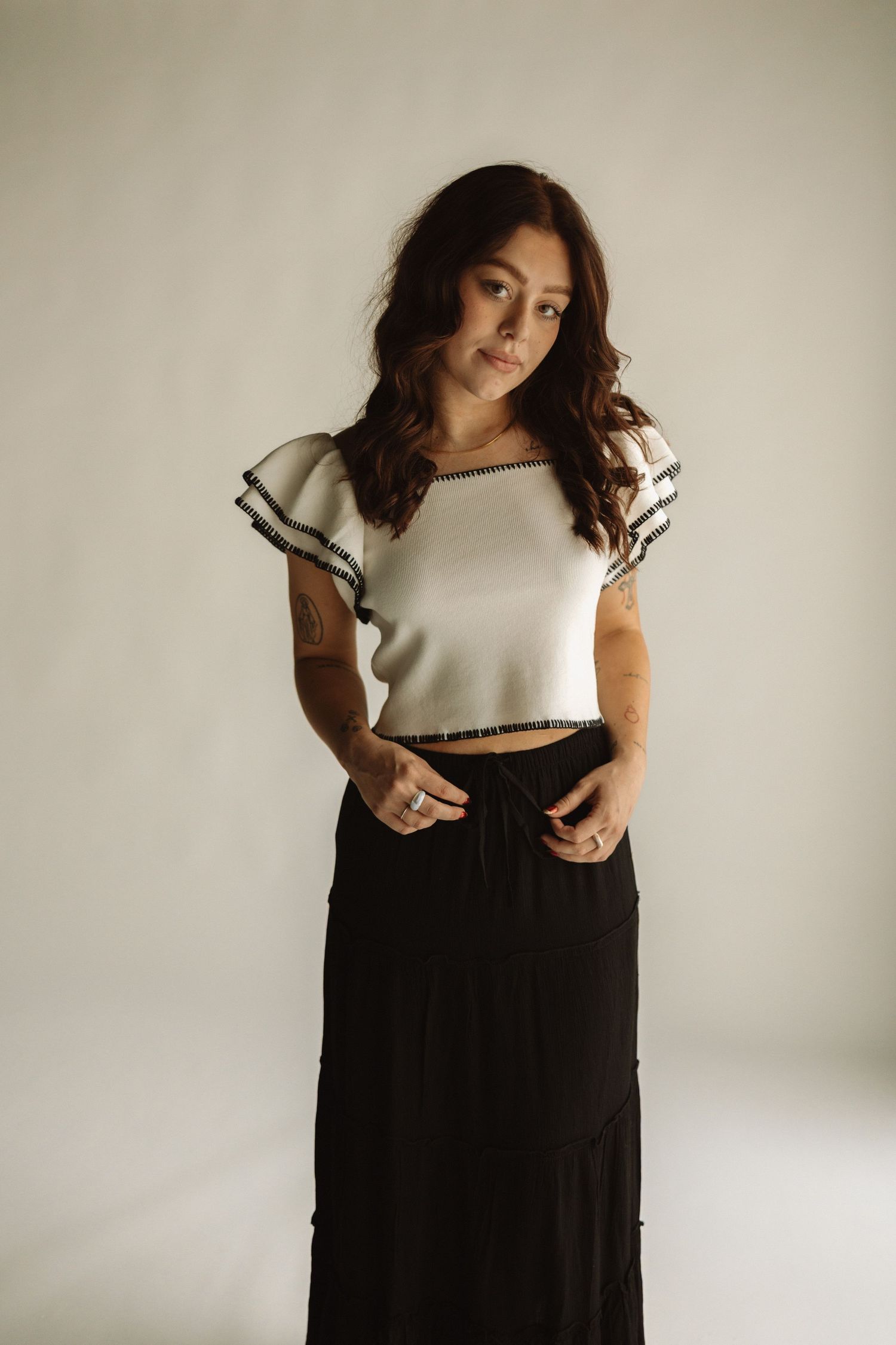 Woman in casual white ruffled crop top and long black skirt poses against neutral studio background in lifestyle portrait.