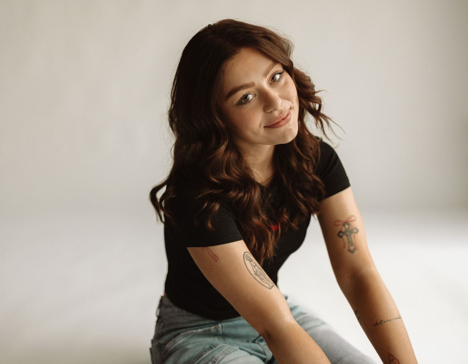 Young woman with long dark wavy hair wearing a black t-shirt and jeans sits casually in a minimalist studio setting.