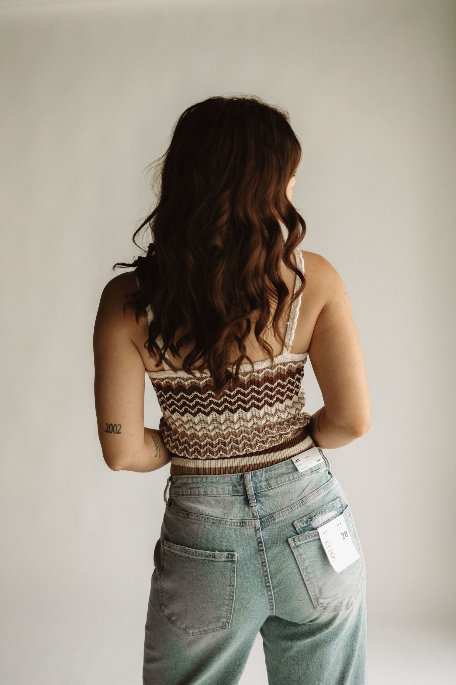Back view of person wearing patterned crop top with wavy brown hair and light wash high waisted jeans against white wall.