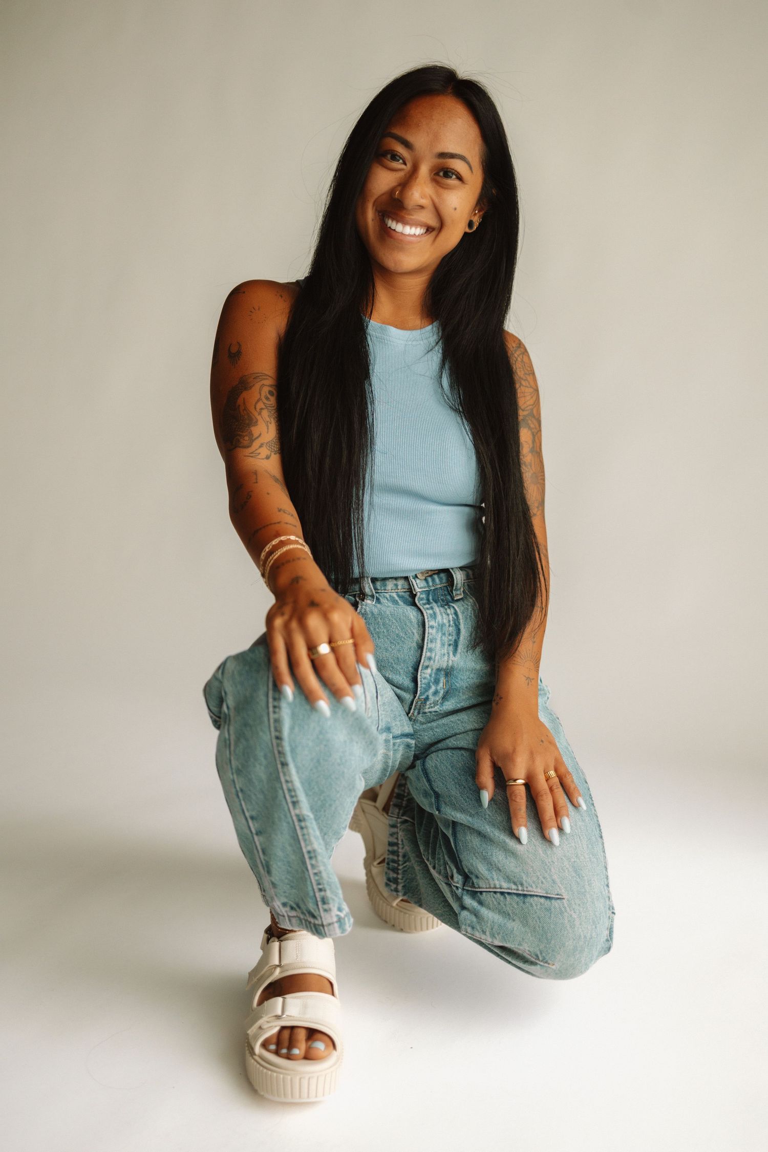 Smiling person in light blue tank top and baggy jeans crouches on white backdrop wearing platform sandals.