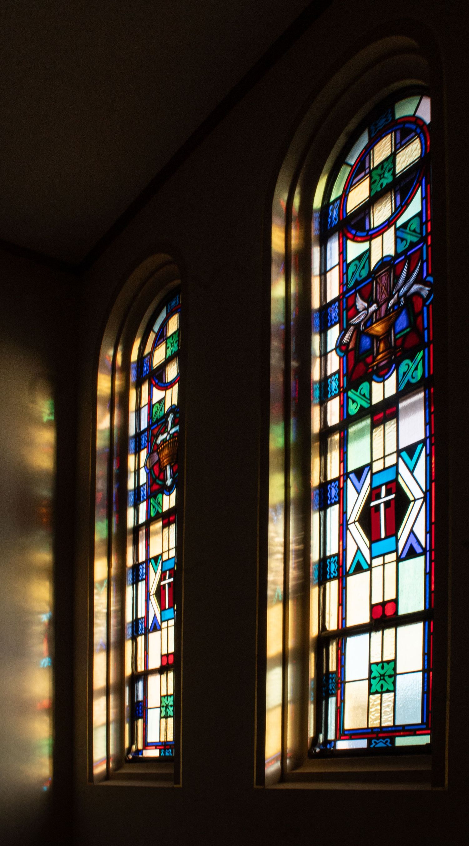 Sunlight streams through a series of colorful stained glass church windows with geometric patterns and religious motifs.
