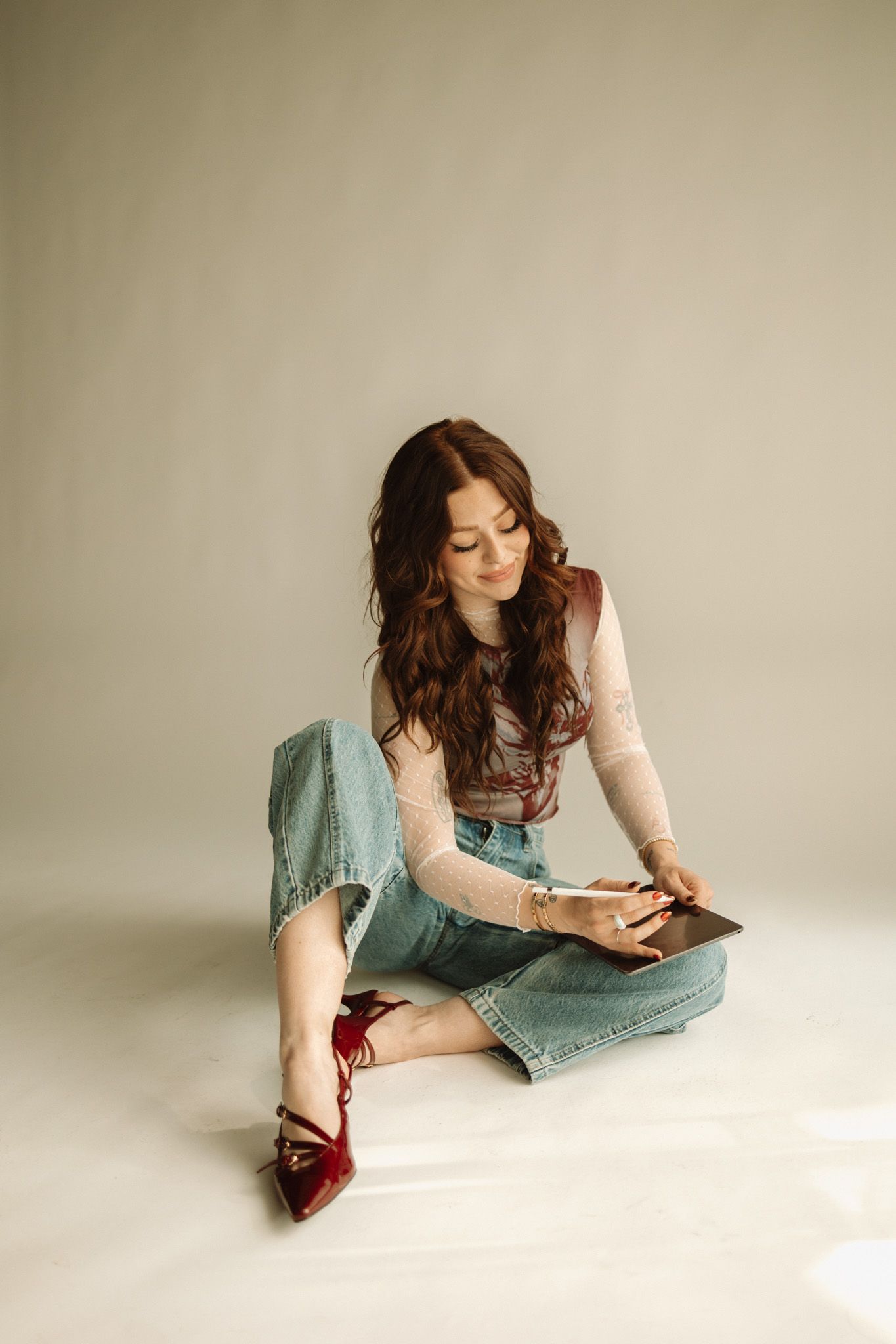 Person in a soft pink sweater and distressed denim overalls reads while sitting on floor wearing burgundy shoes.