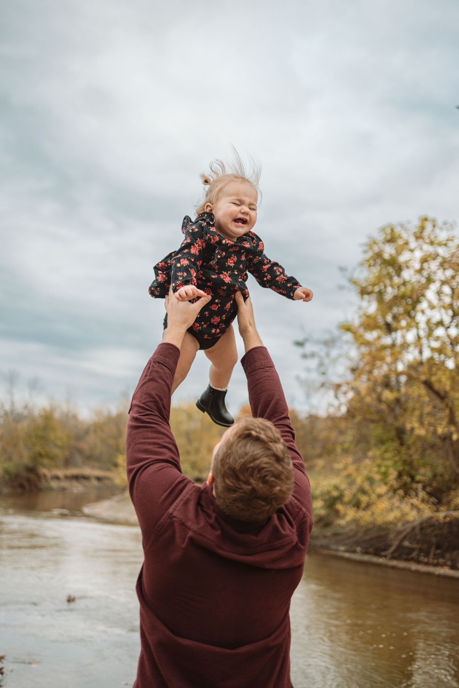 A joyful moment captured by a river as a person in burgundy lifts a laughing child wearing a floral outfit into the air.