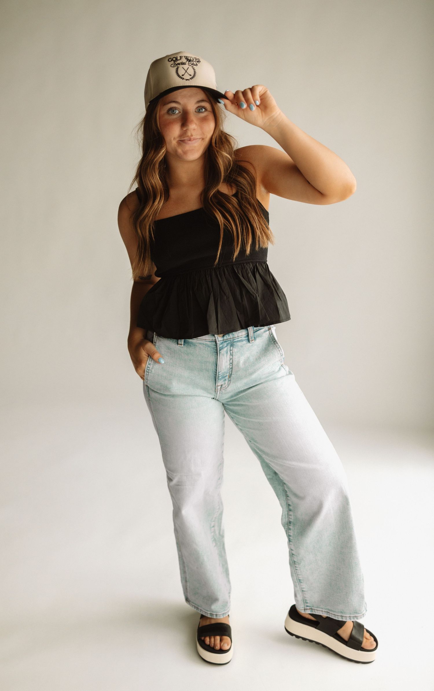 A casual outfit featuring a black peplum top, light wash wide-leg jeans, platform sandals and a baseball cap.