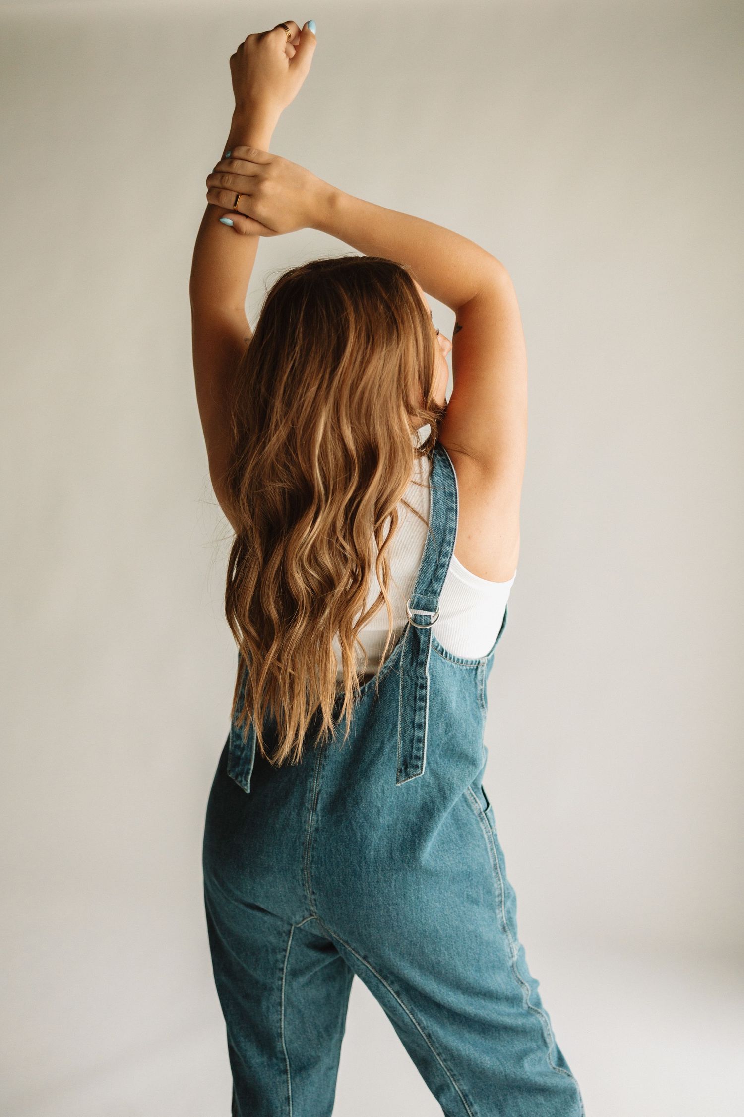 Side view of person in denim overalls tossing their long wavy hair in front of white studio backdrop.