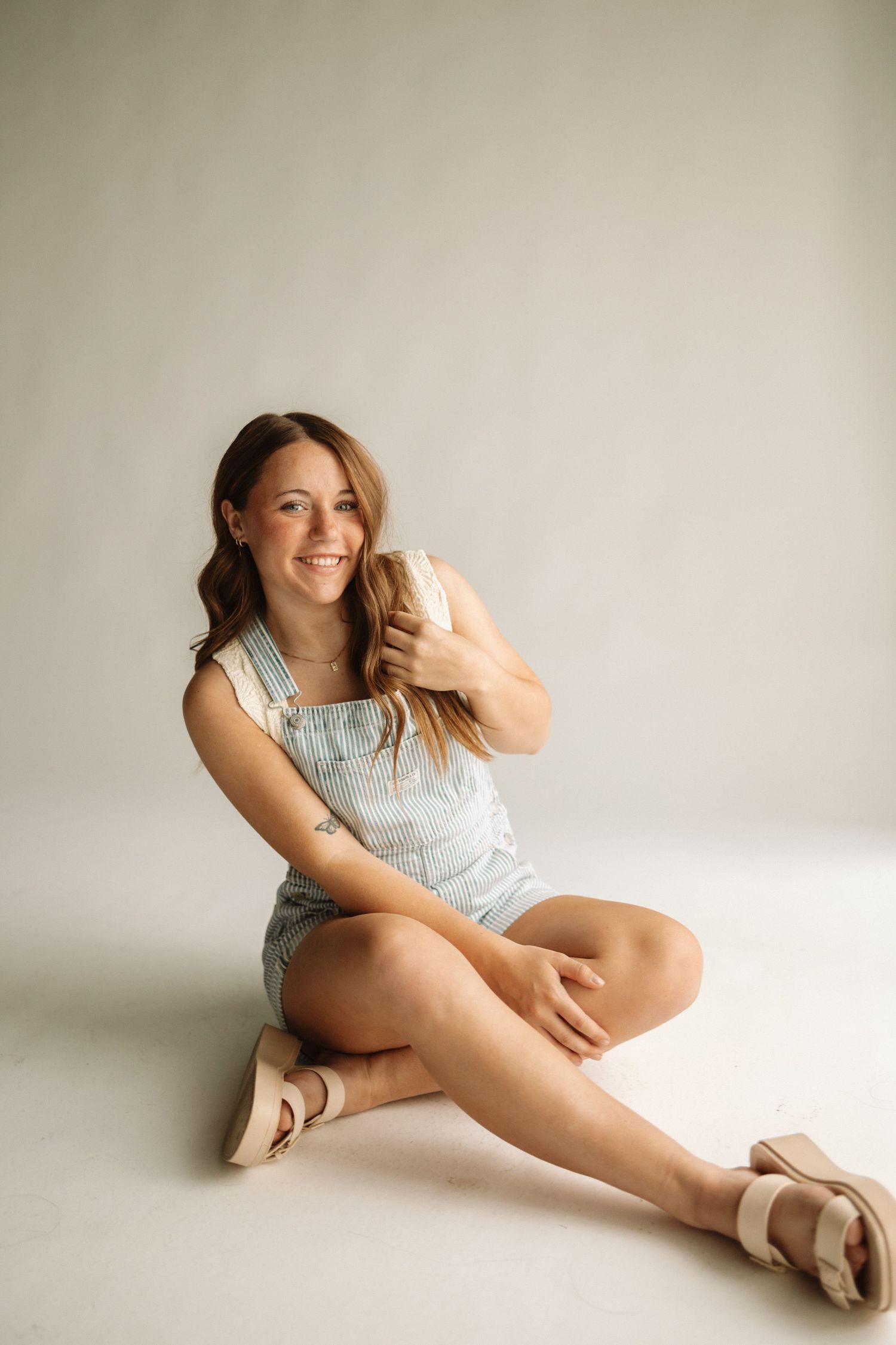 Portrait of someone wearing light blue overalls and ballet flats sitting casually against a neutral studio backdrop.